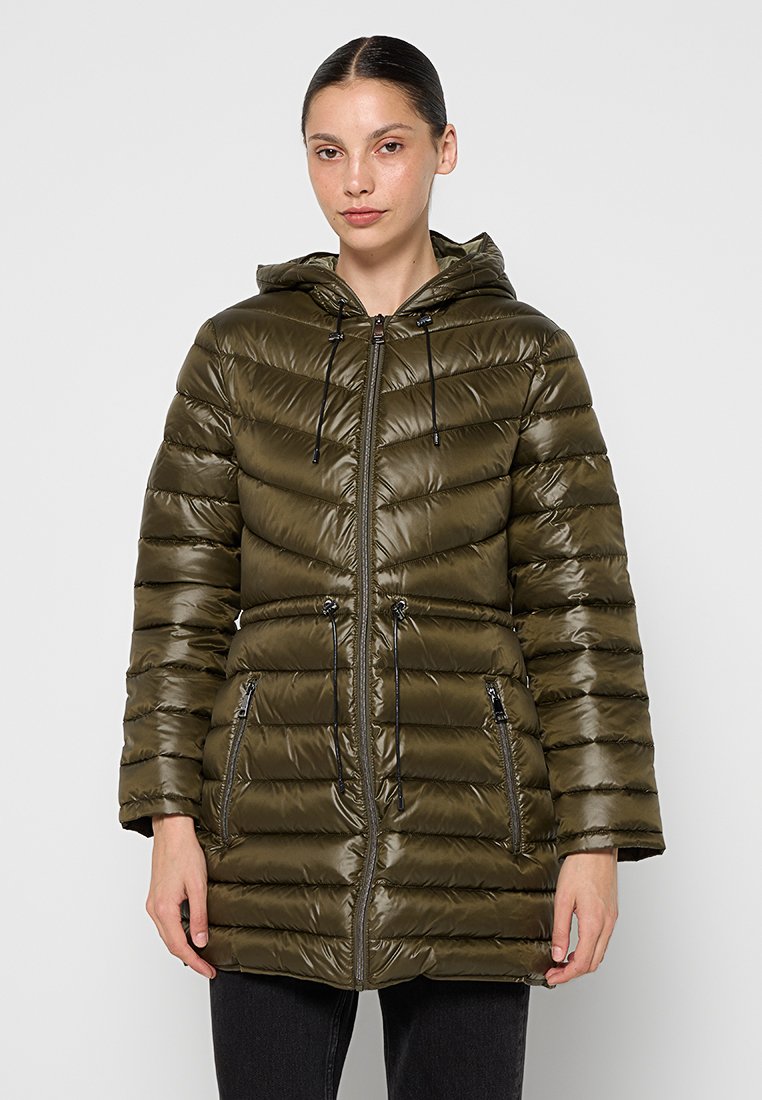 Mujer vistiendo una chaqueta de invierno acolchada con capucha en color verde oliva, con cremallera frontal y cordones ajustables en la cintura, de pie frente a un fondo liso.