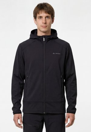 Jonge man die een zwarte, met rits sluitbare capuchonjas draagt met twee voorvakken met rits en het Vaude-logo op de borst, staande tegen een witte achtergrond.