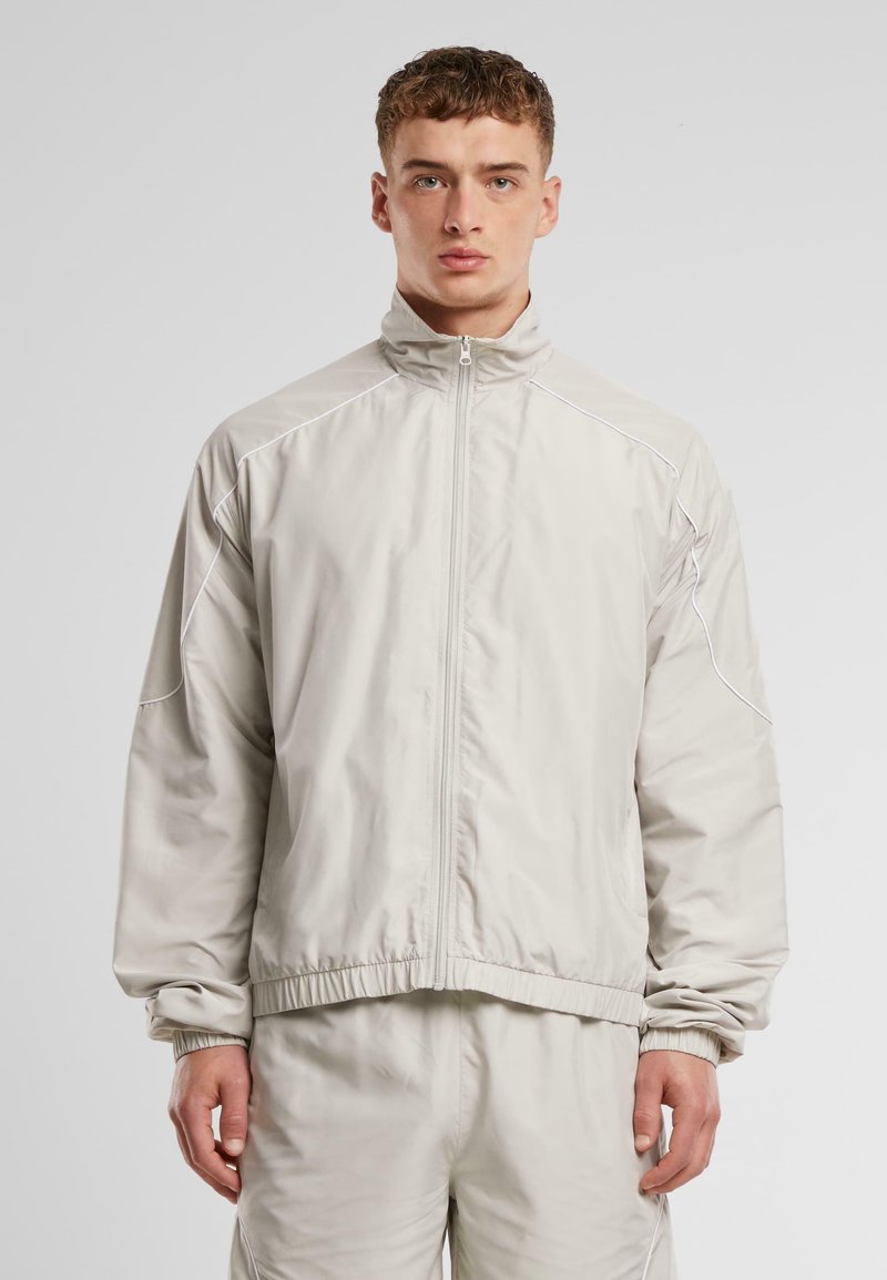 Hellgraue Zip-Jacke mit hohem Kragen, elastischen Bündchen und Saum, mit kontrastierenden weißen Nähten entlang der Schultern und Nähte.
