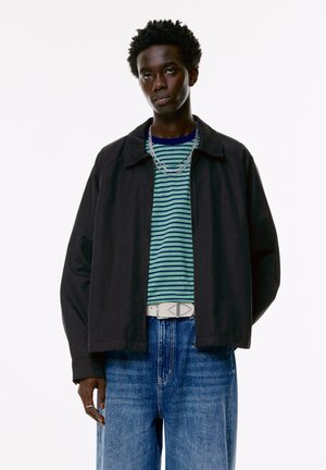 Black collared jacket over a green and blue striped shirt, paired with faded blue jeans and a studded white belt. Silver chain accessories visible.