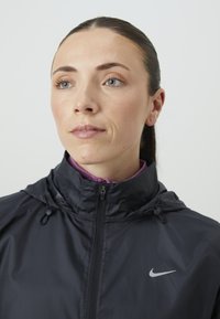 Nike-jakke i sort med høj krave, frontlynlås og hætte med snoretræk. Har en lilla indvendig foring og et sølvlogo på brystet.