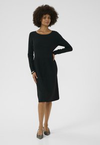 Melna ribota adīta kleita ar garām piedurknēm, apaļu kakla izgriezumu un pieguļošu augstdaļu. Svārki ar vertikālām krokām. Valkāta ar dzīvnieku raksta apaviem.