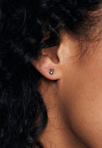 Kolczyki w kolorze złotym z główkami, które zawierają trzy kolorowe kamienie: duży niebieski kamień centralny, otoczony dwoma mniejszymi różowymi kamieniami, osadzone w kwiatowym wzorze.