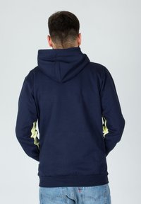 Uomo che indossa un hoodie blu navy con accenti giallo chiaro sulle maniche, in piedi di spalle contro uno sfondo chiaro e semplice.