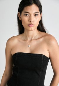 Αμάνικη μαύρη τζιν τοπ με δομημένη εφαρμογή, που διαθέτει ορατές ραφές και ένα ελαφρύ χρυσό κολιέ με μικρό κρεμαστό σε σχήμα καρδιάς.