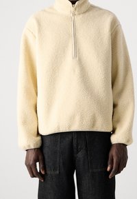 Pullover in pile di un pallido giallo con collo alto e chiusura a mezza zip. Presenta un tessuto testurizzato e una vestibilità rilassata, abbinato a jeans scuri.