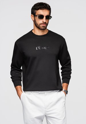 Mężczyzna w czarnej bluzie z napisem, białych spodniach, okularach przeciwsłonecznych i zegarku, stoi z rękami w kieszeniach na szarym tle.