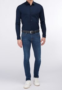 Camisa de botón azul marino con una textura suave, combinada con jeans azul oscuro y zapatillas blancas. Presenta un corte slim y un diseño de cuello clásico.