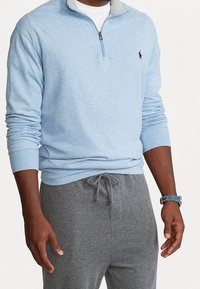 Ljusblå zip-up sweatshirt med grå krage, bomullstyg, långa ärmar och en liten logotyp på bröstet, ihop med gråa svettbyxor.