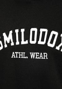 Zwarte sweatshirt met opvallende witte borduring met de tekst "SMILODON" en "ATHL. WEAR." De tekst heeft een textuur en een contrasterend ontwerp.