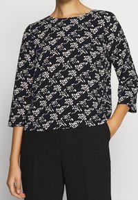 Svart topp med vitt abstrakt mönster, rund halsringning, trekvartsärmar och avslappnad passform, tillverkad av lättviktsmaterial.