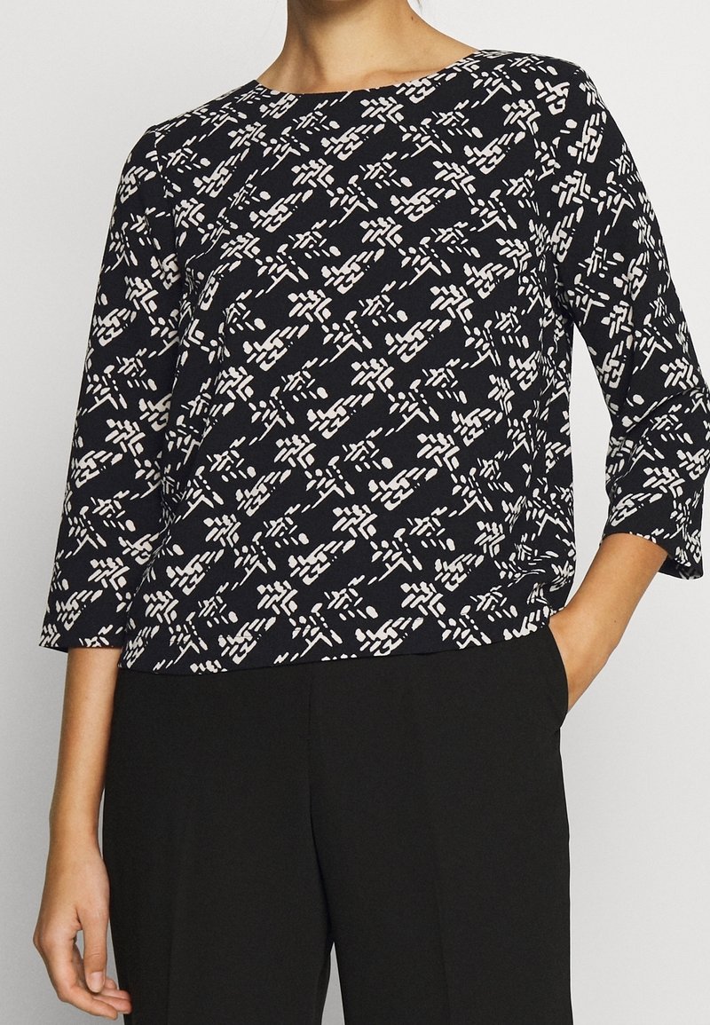 Svart topp med vitt abstrakt mönster, rund halsringning, trekvartsärmar och avslappnad passform, tillverkad av lättviktsmaterial.