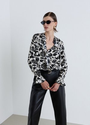 Donna con capelli corti che indossa occhiali da sole neri, camicetta floreale bianca e nera, pantaloni di pelle neri, orecchini e anelli d’oro, in piedi al chiuso.