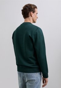 Bărbat cu părul scurt, brunet, îmbrăcat într-o cămașă cu mânecă lungă, verde închis și blugi bleu deschis, întors de profil în fața unui fundal simplu, gri deschis.