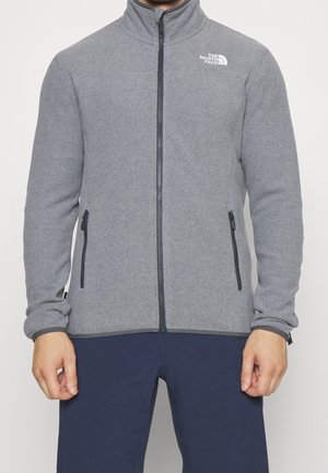 Mężczyzna noszący szarą bluzę polarową na zamek błyskawiczny z czarnymi zamkami i logo The North Face na piersi, w połączeniu z ciemnoniebieskimi spodniami.