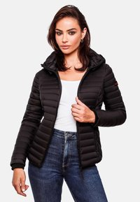 Veste matelassée noire avec capuche, design matelassé, silhouette ajustée, fermeture éclair et poignets côtelés. Associée à un haut blanc et un jean en denim.