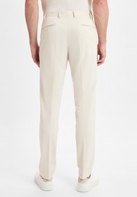 Beige Tailored-Hosen aus leichtem Stoff, mit zwei Gesäßtaschen mit Knopfverschluss und glatter Textur. Die Schuhe sind weiß.