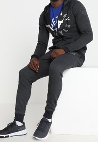Hombre sentado sobre un bloque blanco, vistiendo una sudadera con cremallera gris oscuro, pantalones jogger a juego, camiseta gráfica azul, calcetines blancos y zapatillas negras.