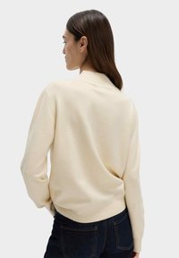 Suéter color crema con un acabado suave y texturizado, con mangas largas y un dobladillo redondeado, combinado con jeans de mezclilla oscura.