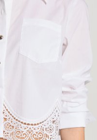Weißes Hemd mit Knopfleiste und einer einzelnen Tasche, verziert mit detaillierter Spitzenborte am Saum. Der Stoff wirkt glatt, mit leicht gerafften Ärmeln.