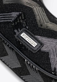 Černá taška se flitry, která má texturovaný vzor s kovovými akcenty a kožený štítek se značkou. Kompaktní design s elegantním provedením.