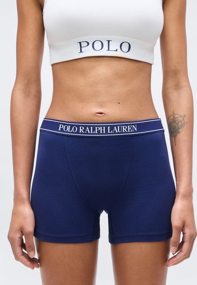 Γυναίκα με μπλε σκούρα boxer briefs Polo Ralph Lauren και λευκό αθλητικό σουτιέν με την επιγραφή "POLO", στέκεται με ορατό τον κορμό και τατουάζ στο μπράτσο.