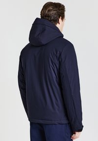 Veste isolante bleu marine avec capuche, présentant une texture lisse et des poignets ajustables. Design simple sans motifs ni quincaillerie visibles.