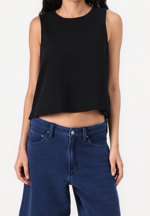 Frau trägt ärmelloses schwarzes kurzes Top und hoch taillierte blaue Jeans, steht mit entspannt an den Seiten hängenden Armen.