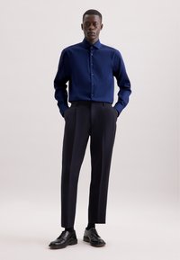 Marineblauwe overhemd met knopen en een gestructureerde pasvorm, gecombineerd met op maat gemaakte zwarte broeken en zwarte leren schoenen, tegen een neutrale achtergrond.