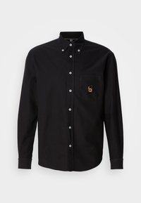 Camisa negra de manga larga con botón, cuello tipo button-down y un bolsillo en el pecho que presenta un pequeño logo bordado en oro y negro.