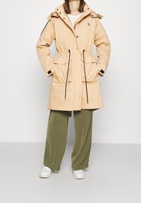 Beige, waterproof parka with a drawstring waist, two large front pockets, a detachable hood, and black accents along the zippers and sleeves.