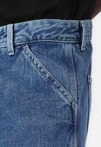 Jean en denim d'un bleu moyen, avec une taille, des passants de ceinture et des poches avant diagonales ; texture lisse et détails de couture.