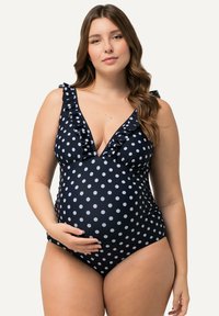 Costume da bagno premaman in blu navy con pois bianchi, caratterizzato da bordo arricciato lungo il scollo e un profondo scollo a V, realizzato in tessuto elasticizzato.