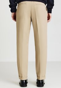 Pantaloni beige su misura con una texture liscia, dotati di due tasche posteriori e orlo arrotolato, abbinati a scarpe nere.