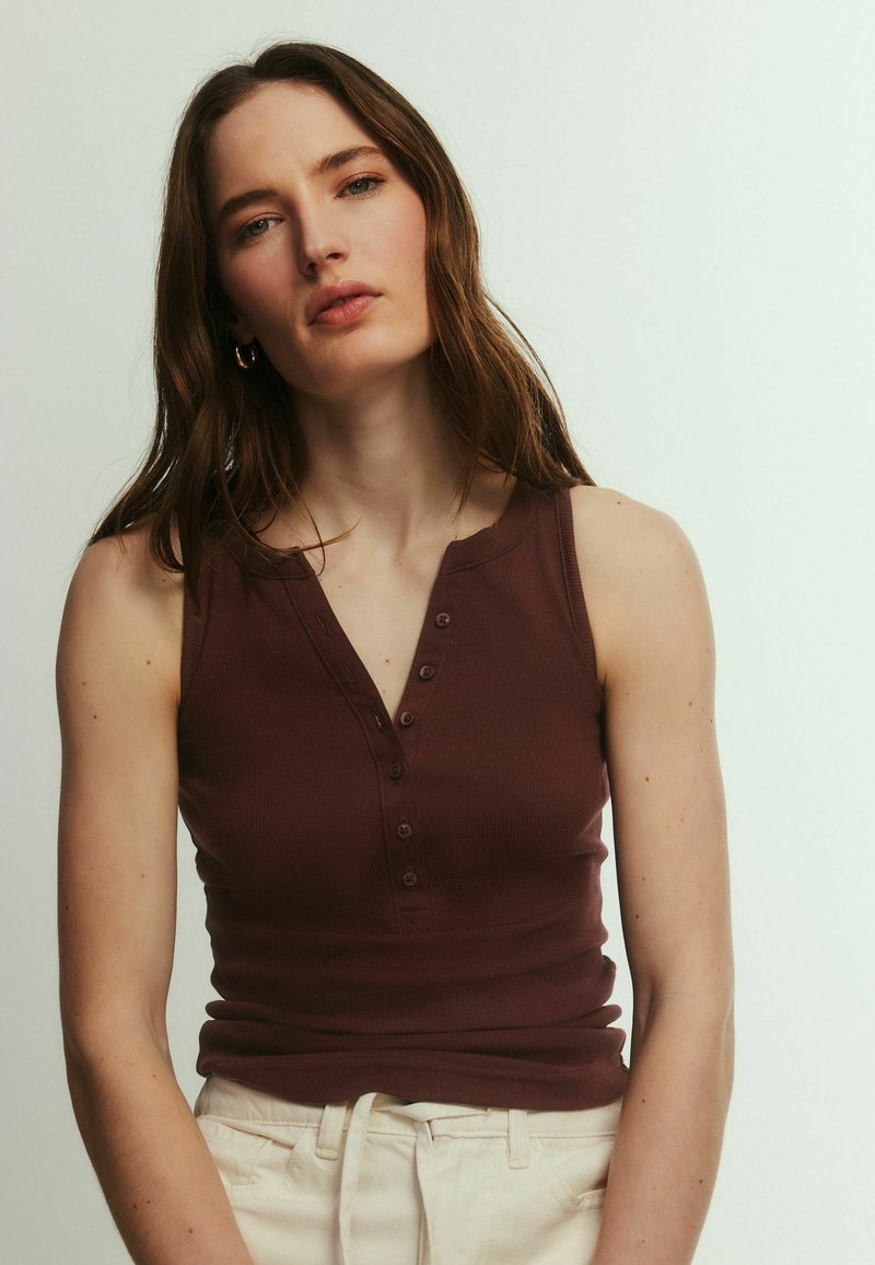 Joven mujer con pelo largo y castaño, vestida con una blusa de botones sin mangas de color marrón oscuro y pantalones de color claro sobre un fondo liso.