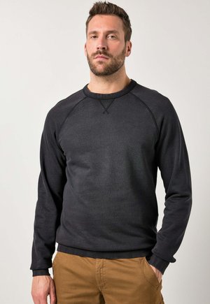 Mann mit Bart, der einen schwarzen Langarmpullover und braune Hosen trägt, steht mit einer Hand in der Tasche vor einem schlichten, hellen Hintergrund.