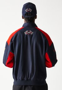 Μπλε ναυτικό και κόκκινο μπουφάν με γιακά, με λογότυπα στο πίσω μέρος. Λείο ύφασμα και σχεδιασμός με ελαστική μέση.