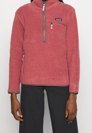 Person iført en støvet rosa Patagonia fleecepullover med kvartlynlås og lynlåslomme på brystet, kombineret med sorte bukser.