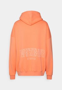 Orange hoodie med avslappnad passform. Har stor vit text "WRSTBHNSTUDIO" tryckt på baksidan. Mjuk tyg, ribbade muddar.