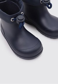 Paire de bottes de pluie en caoutchouc bleu marine avec fermetures réglables par cordon et semelles texturées sur une surface gris clair.