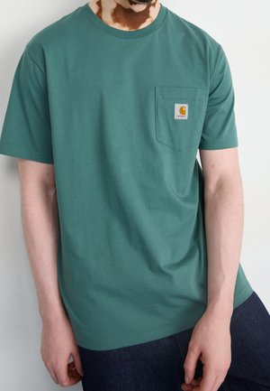 Homme portant un t-shirt vert à manches courtes avec une poche poitrine et un patch logo Carhartt, associé à un jean bleu foncé.