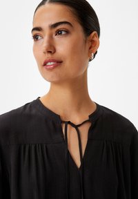 Schwarze Bluse mit rundem Ausschnitt, vorderem Bindedetail und subtiler Textur. Verfügt über sanft gerafftes Material entlang der Schultern und des Knopfleisten.