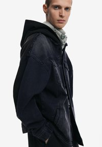 Mörk denimjacka med kontrasterande svart huva, fransade kanter och framfickor. Ljust grå mönstrad bandana som bärs under jackan.