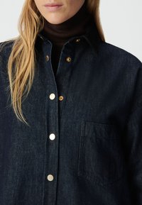 Zbliżenie osoby w ciemnoniebieskiej kurtce dżinsowej ze złotymi guzikami, noszącej brązowy sweter z golfem, ukazujące długie blond włosy i dolną część twarzy.