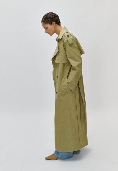 Γυναίκα με μακρύ χακί παλτό τύπου τρανς με φαρδιές πέτσωτες, μπλε τζιν, καφέ μυτερά παπούτσια, στέκεται πλευρικά μπροστά σε απλό λευκό φόντο.