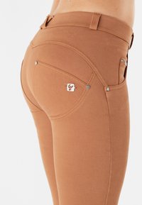 Jeans in denim marrone con un design aderente, tasche posteriori curve distintive e bottoni metallici in evidenza. Tessuto liscio e senza ulteriori fantasie.