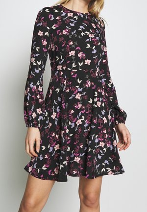 Kvinde iført en sort langærmet kjole med rosa, lilla og hvide blomstermønstre og en flagrende nederdel foran en ensfarvet baggrund.