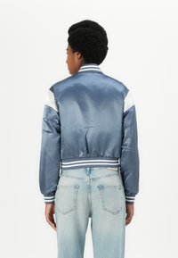 Jachetă bomber din satin albastru, cu manșete și tiv din material elastic, având accente albe pe umeri și un design ajustat. Asortată cu blugi de culoare albastră deschisă.