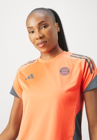 Πορτοκαλί αθλητικό μπλουζάκι με γκρι λεπτομέρειες, κοντά μανίκια και στρογγυλή λαιμόκοψη. Διαθέτει το λογότυπο της FC Bayern München στο μπροστινό μέρος.