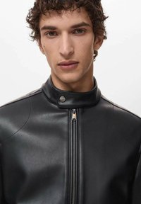 Giubbotto di pelle nera con colletto alto, dotato di chiusura con zip e dettagli sottili sulle cuciture delle spalle. Texture liscia e design aderente.